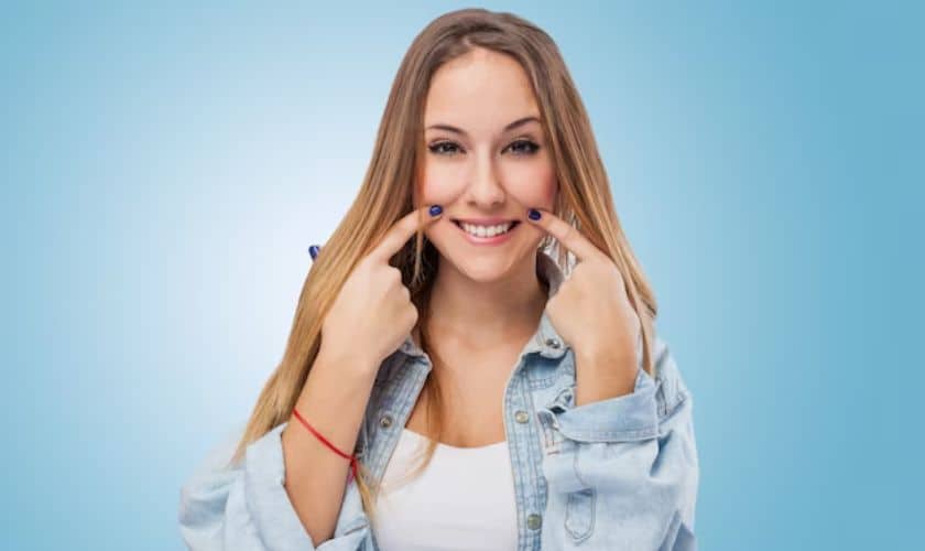 Smiling woman in denim shirt pointing to her teeth against blue background for orthodontics clinic