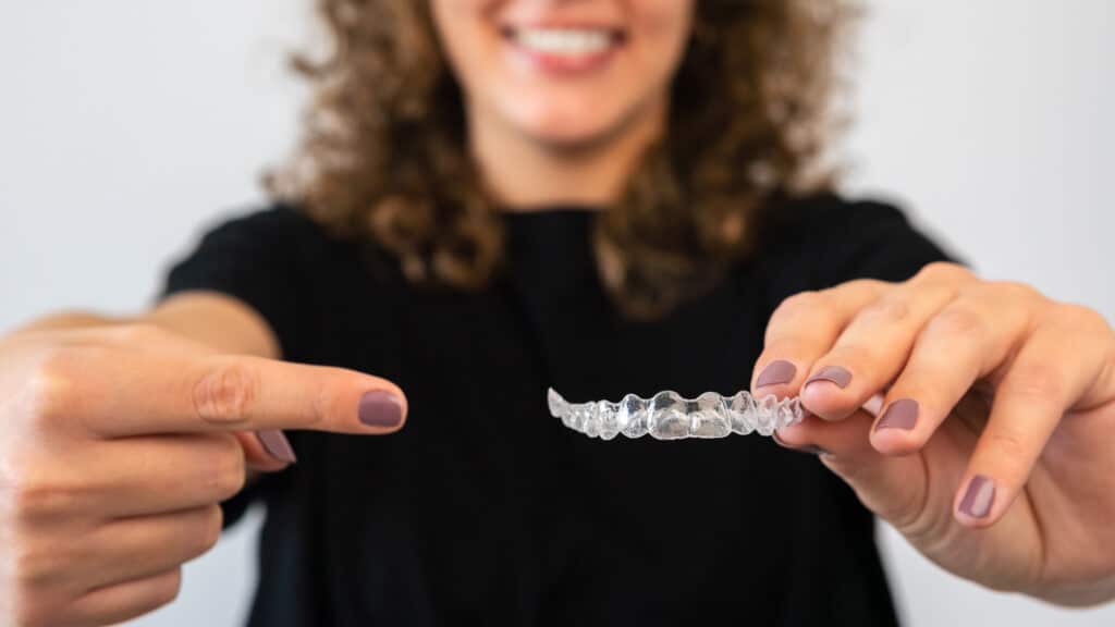 A smiling person holds a clear dental aligner with both hands. The focus is on the aligner; the person's face is slightly blurred, conveying a positive tone.